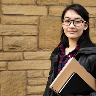 An international student wearing glasses and holding books stood in front of a brick wall