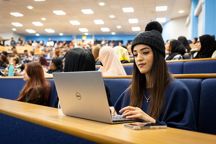 Student wearing a hat and working on a laptop in a lecture hall