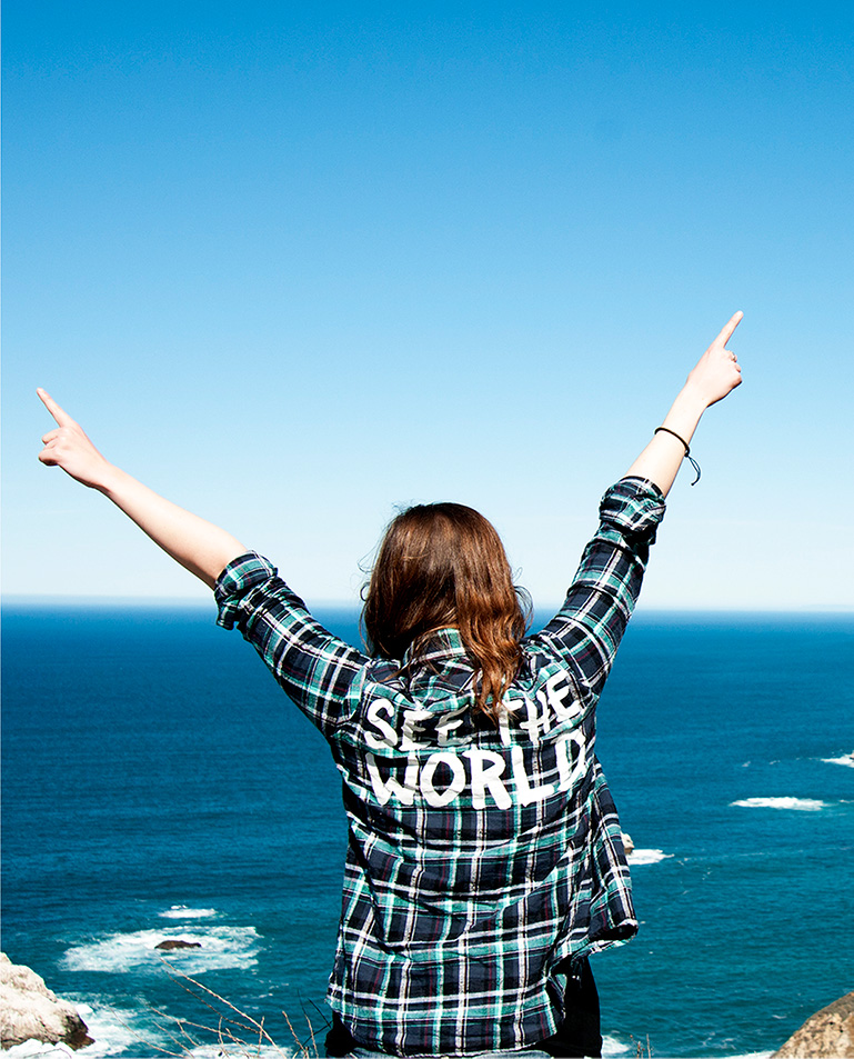 Student with back towards the camera, arms spread, overlooking a sea. The student is wearing a shirt saying 'See the world'.