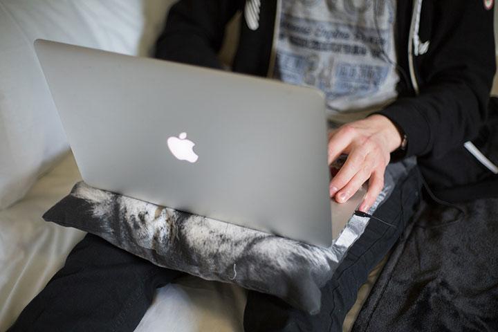 A close-up of the back of a laptop rested in a student's lap. One of the student's hands is visible, typing on the keyboard.