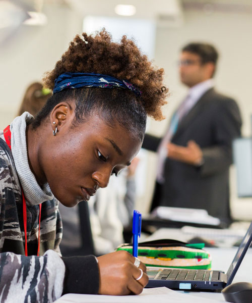 A student writing at a desk. A lecturer is in the background.